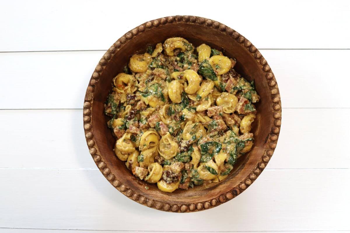 Bowl of tortellini pasta salad with spinach, diced salami, and sun-dried tomatoes on white wooden surface.