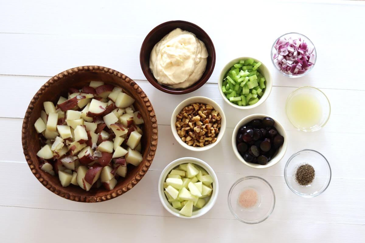Bowls of chopped potatoes, apple, celery, walnuts, grapes, onion, and dressing ingredients arranged on table.