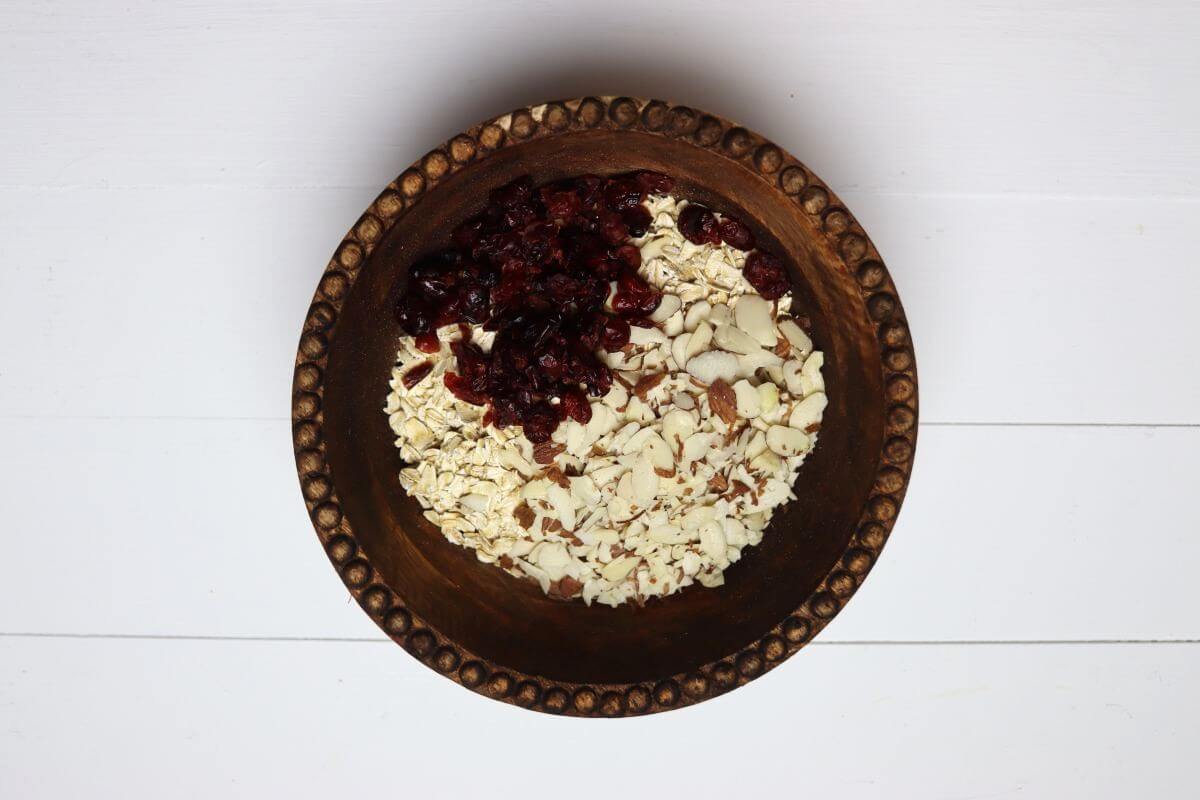 Oats sliced almonds and dried cranberries in wooden bowl on white surface.