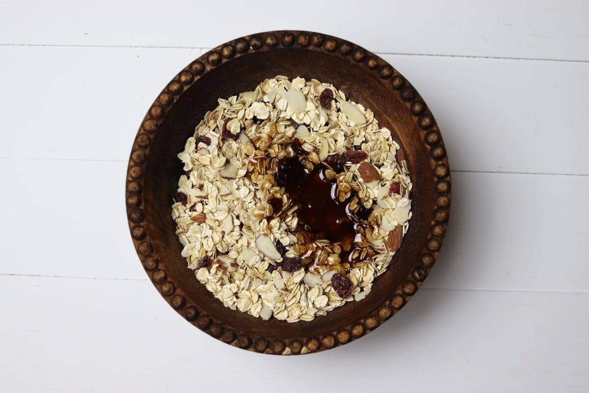 Oats almonds cranberries and honey mixture in wooden bowl on white surface.