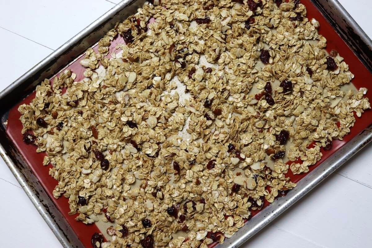 Oat mixture with almonds and cranberries spread on silicone-lined baking sheet.