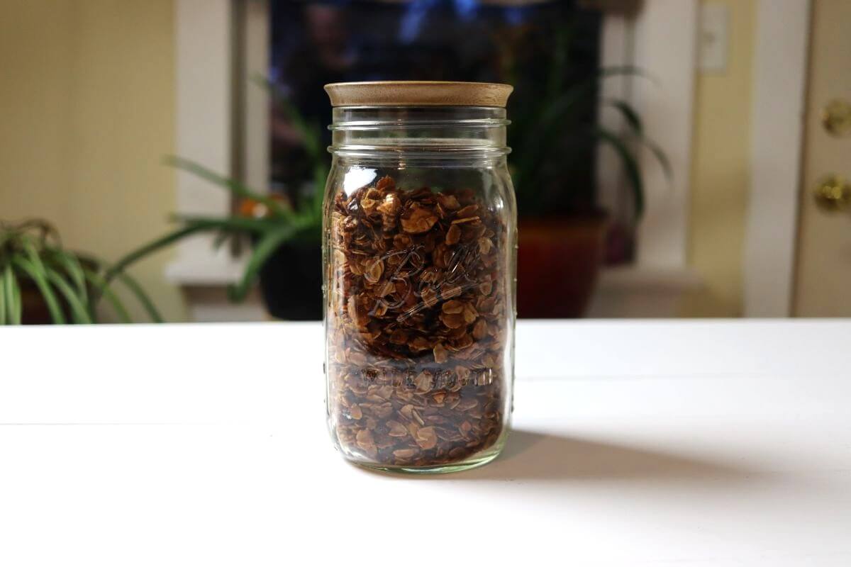 Glass jar filled with baked granola on white surface with blurred kitchen background.