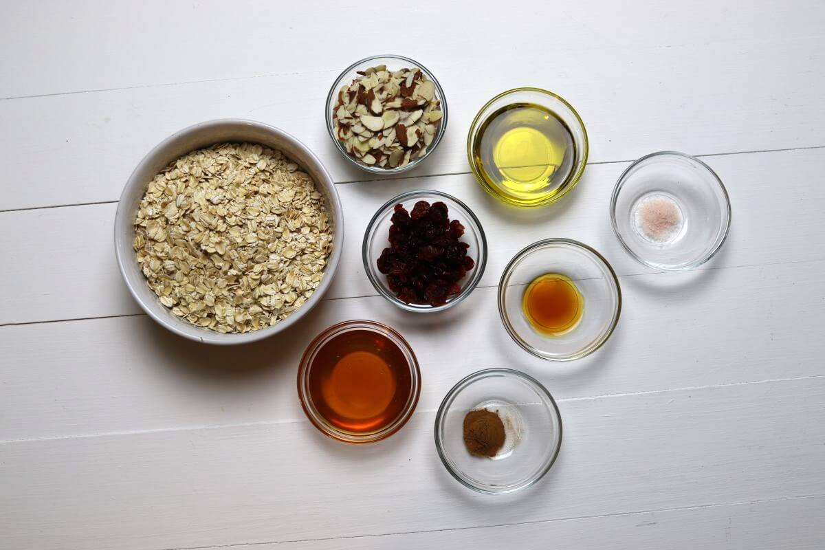 Bowls of oats almonds cranberries honey oil vanilla salt and cinnamon on table.