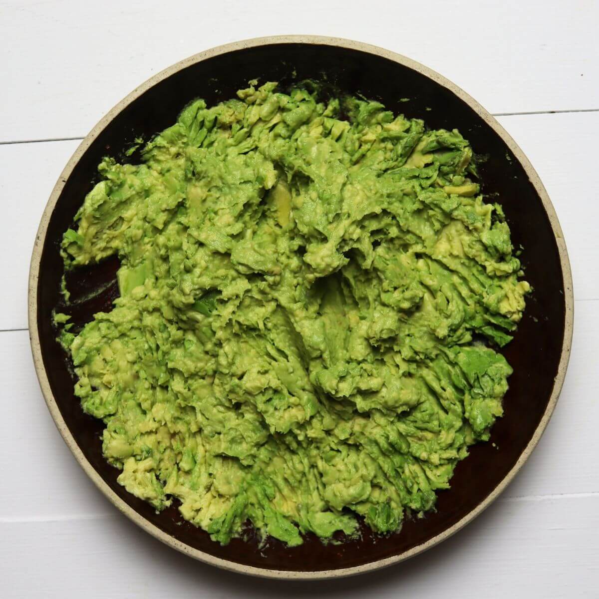 Mashed avocado in a bowl with a coarse texture on a white surface.
