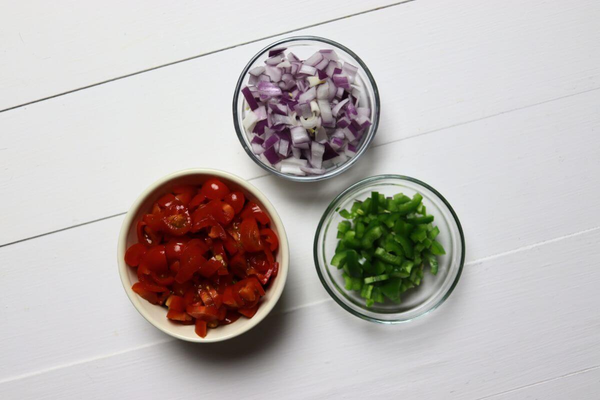 Bowls of diced grape tomatoes, red onion, and jalapeño on a white surface.