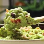 Spoon lifting guacamole with tomato and onion pieces above a bowl with text overlay “Fresh guacamole from scratch Keep it simple. Keep it fresh".
