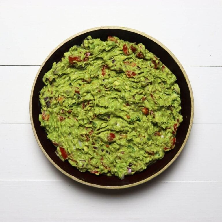 Bowl filled with guacamole showing chunks of tomato and red onion on a white surface.