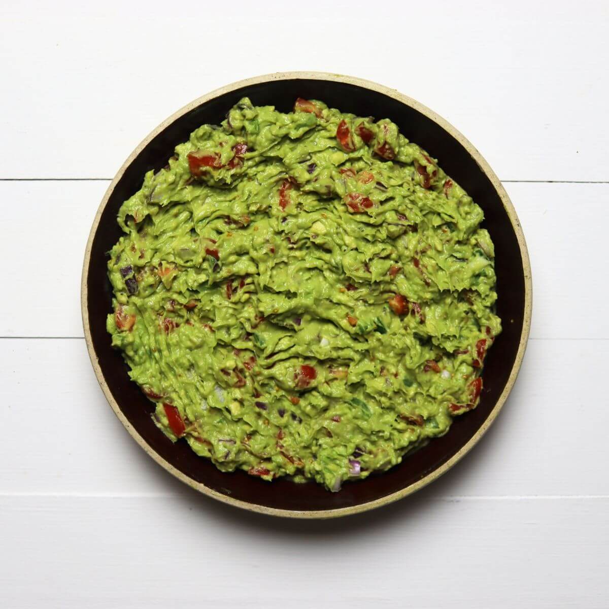 Bowl filled with guacamole showing chunks of tomato and red onion on a white surface.