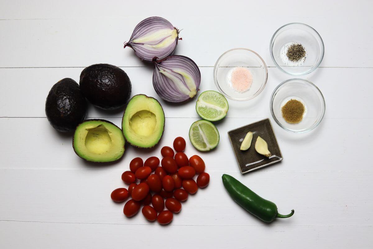 Avocados, red onion halves, grape tomatoes, jalapeño, lime, garlic, and small bowls of spices on a white surface.