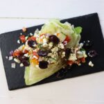 Iceberg lettuce wedge topped with chopped vegetables, feta crumbles, olives, and vinaigrette.
