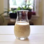 Glass carafe of lemon oregano vinaigrette on white table with blurred plants in background.