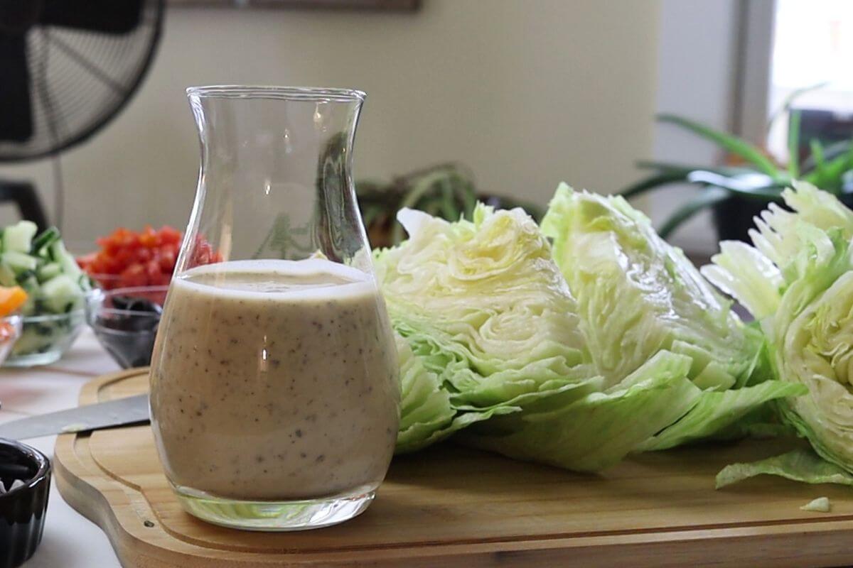 Glass jar of lemon oregano vinaigrette on board with iceberg lettuce wedges and chopped vegetables.