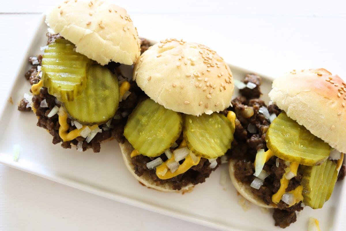 Three loose meat sliders topped with pickle slices, mustard, and chopped onion on plate.
