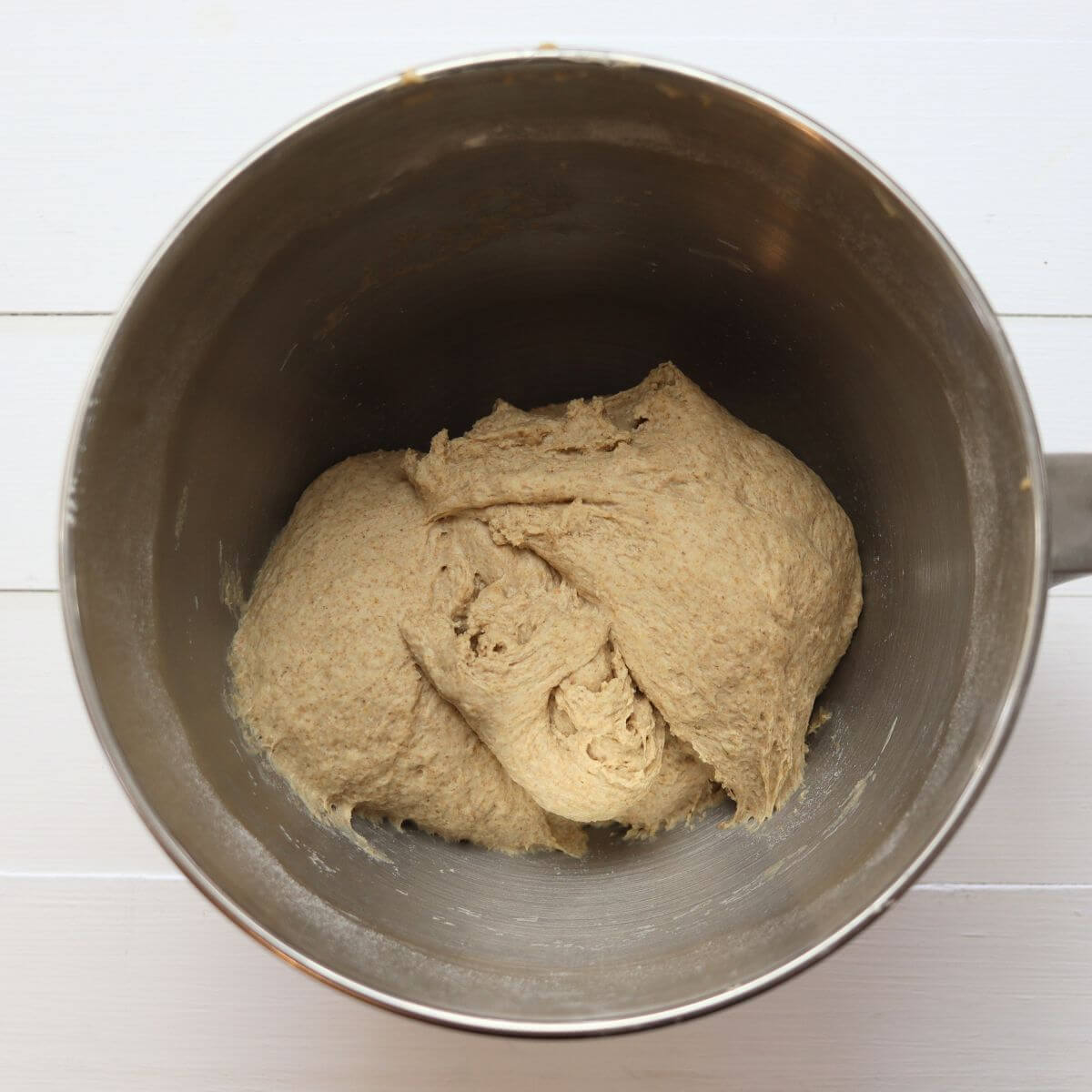 Kneaded whole wheat bread dough inside stand mixer bowl on white surface.