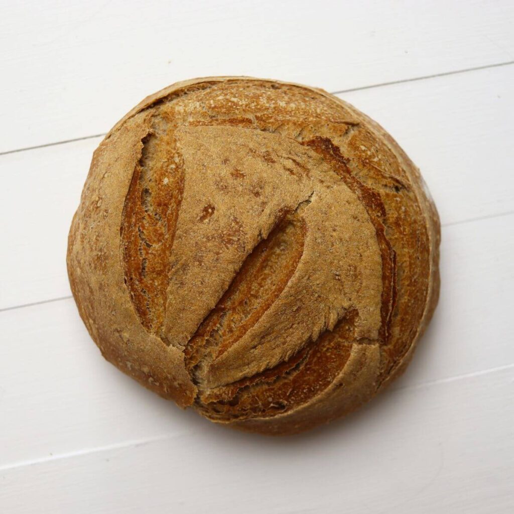 Round whole wheat bread loaf showing split crust and scoring on white surface.