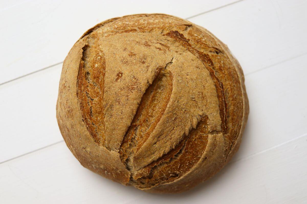 Rustic whole wheat bread loaf with scored top placed on white surface.