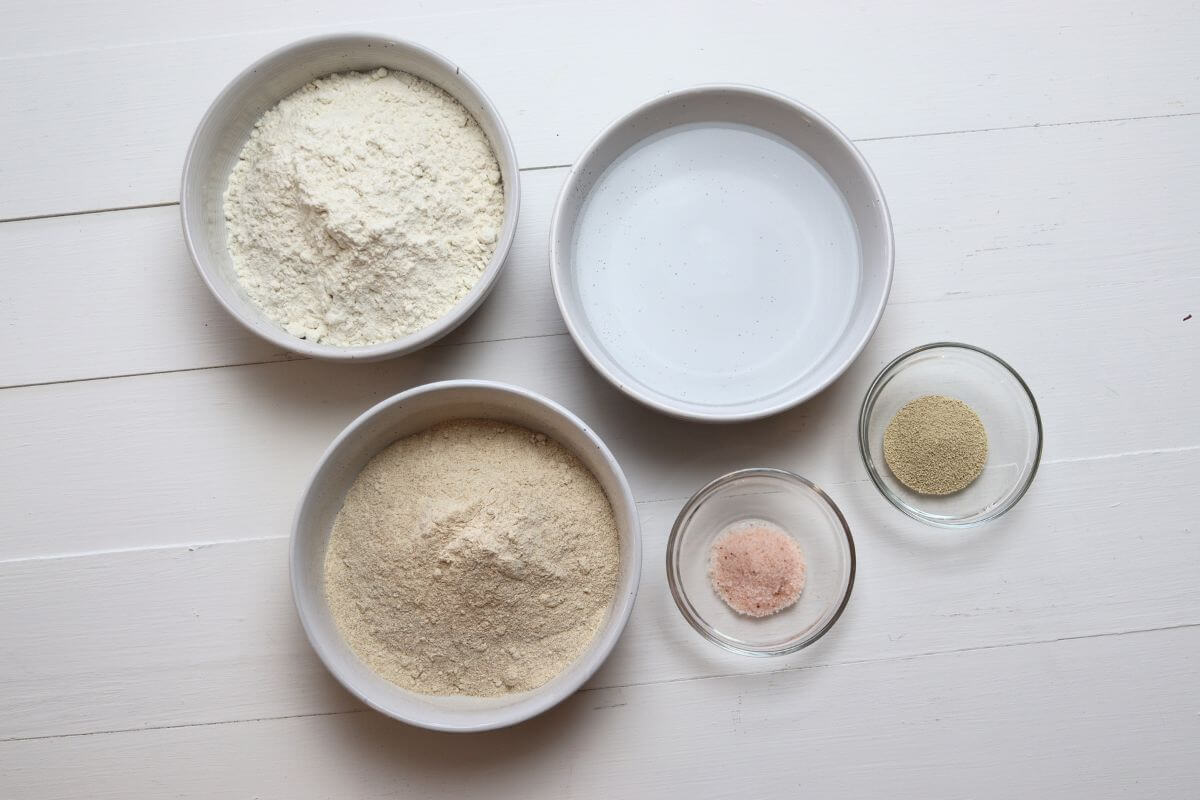 Bowls of whole wheat flour, all-purpose flour, water, yeast, and salt on white surface.