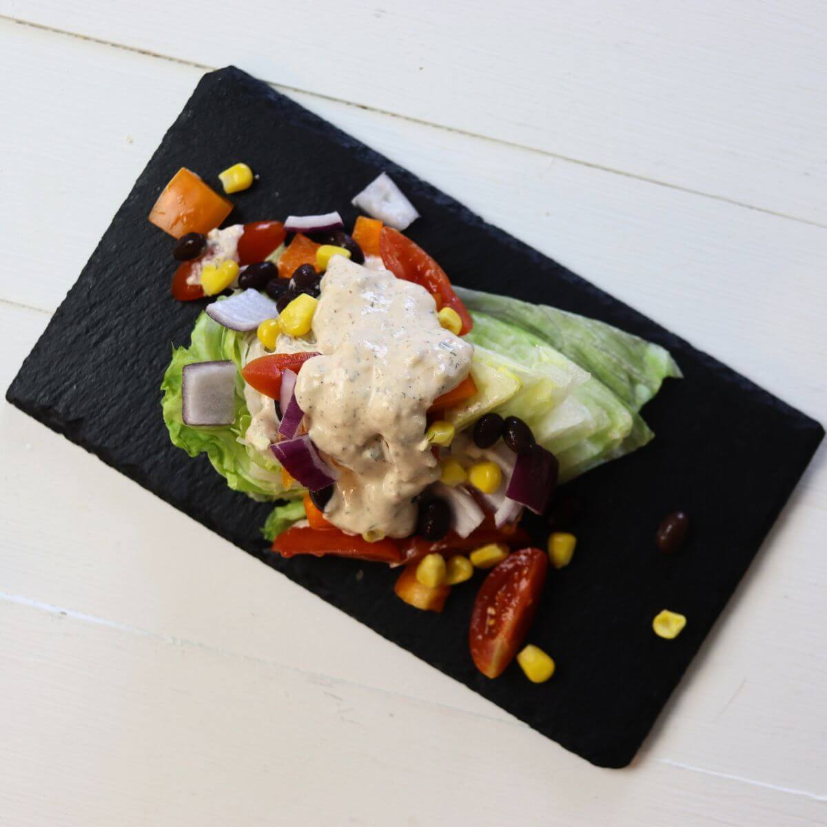 Iceberg wedge topped with black beans, corn, tomatoes, onion, and creamy dressing on slate plate.