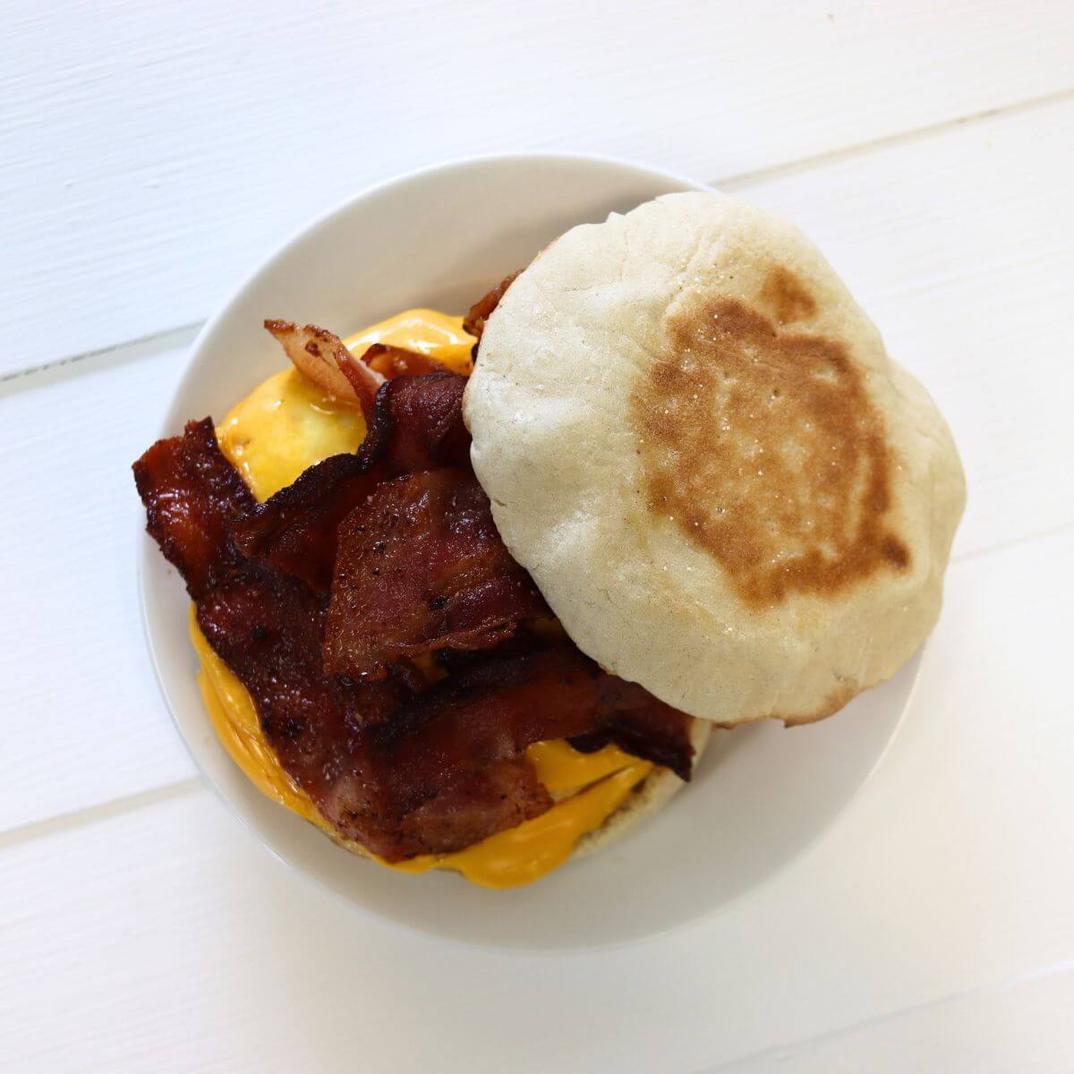 English muffin sandwich with bacon strips and melted cheese on a white plate.