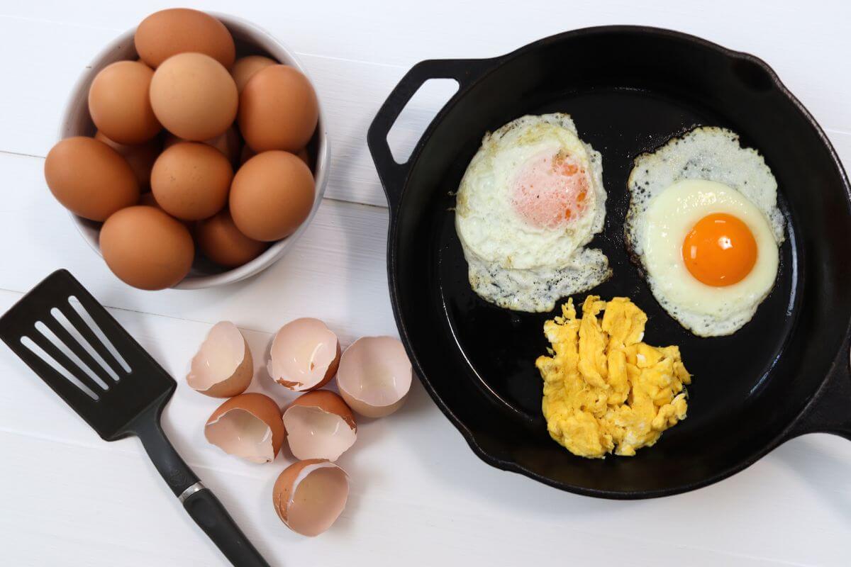 Cast iron skillet with fried eggs, scrambled eggs, eggshells, spatula, and bowl of eggs.