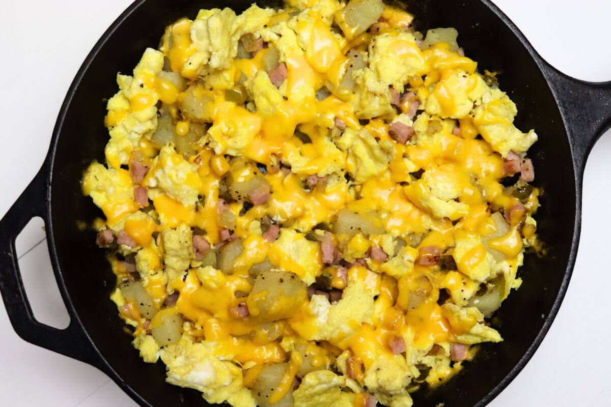 Cast iron skillet filled with scrambled eggs, diced ham, potatoes, and melted cheddar cheese.