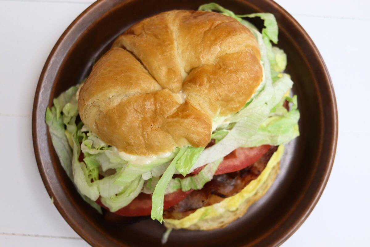 Egg BLT croissant sandwich with lettuce, tomato, bacon, and folded egg on a brown plate.