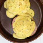 Eggs Benedict with Canadian bacon and hollandaise sauce on a brown plate with “Eggs Benedict with From Scratch Hollandaise” text overlay.