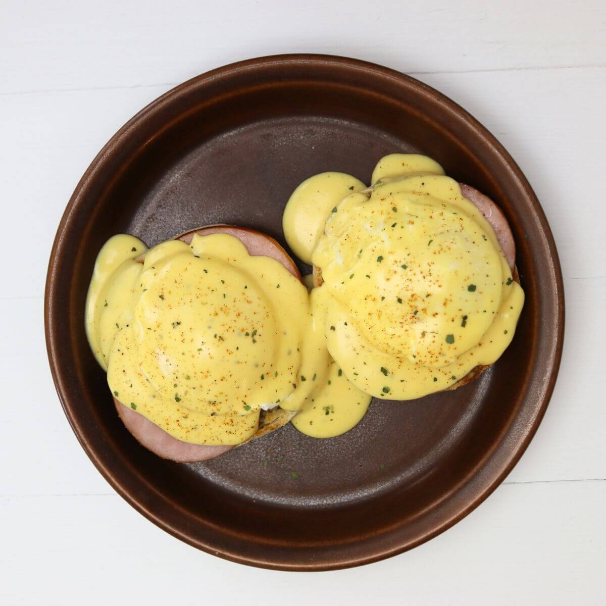 Eggs Benedict topped with hollandaise sauce and seasoning on a brown plate.