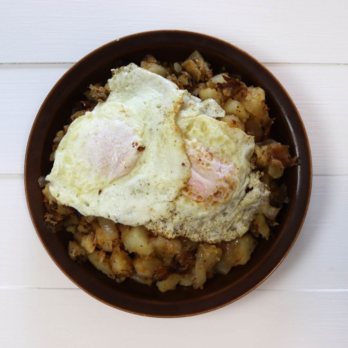 Brown bowl filled with home fries and topped with two cooked eggs.