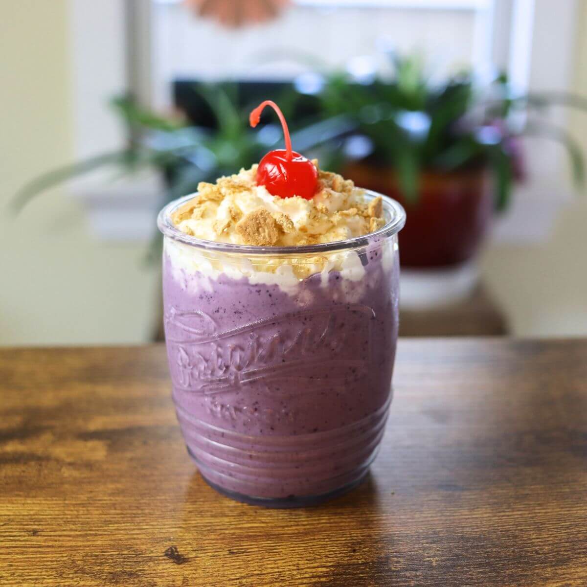 Glass jar filled with a purple milkshake and topped with whipped cream, crumbs, and a cherry.