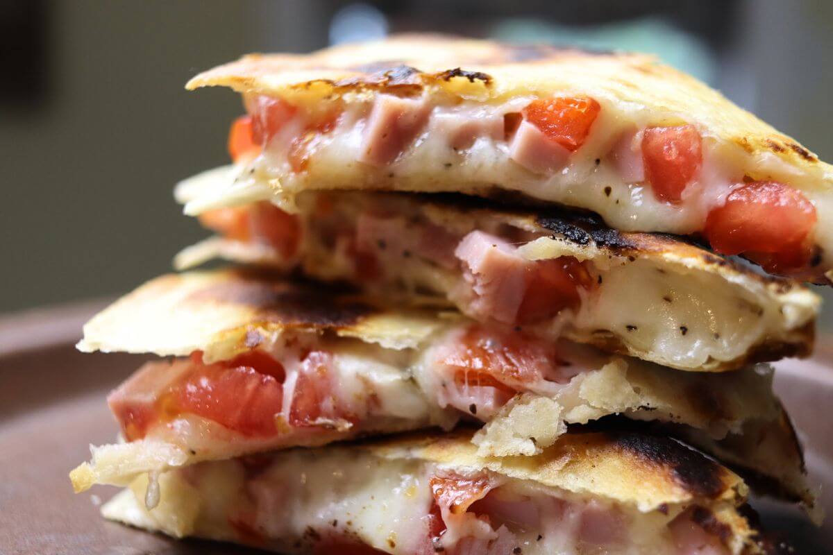 Grilled ham and cheese quesadilla wedges stacked on a plate with melted cheese and diced tomatoes.