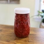 Jar of red fruit jam with a white lid on a table.