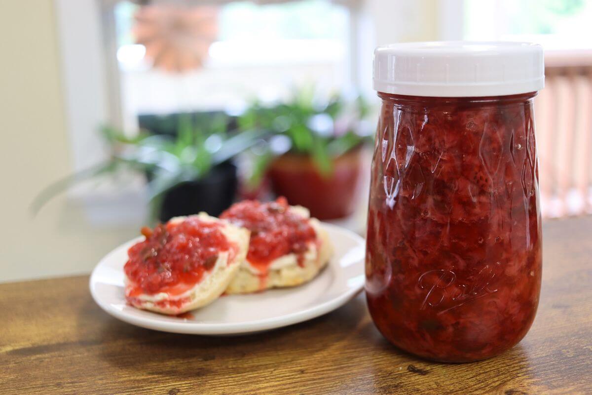 Jar of red jam beside two biscuit halves topped with jam on a white plate.