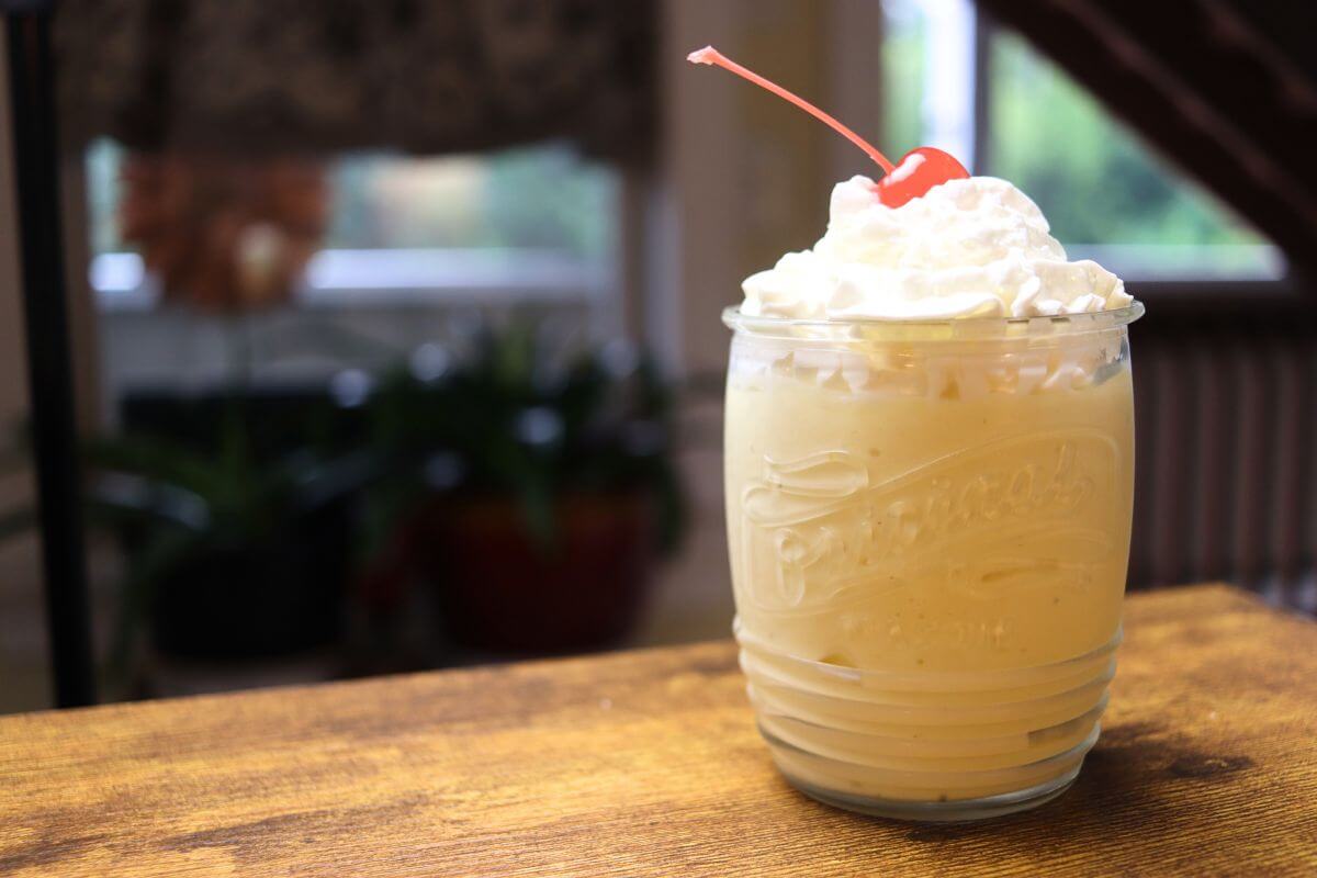 Glass jar filled with a tropical fruit milkshake, whipped cream, and a maraschino cherry on a wooden table.