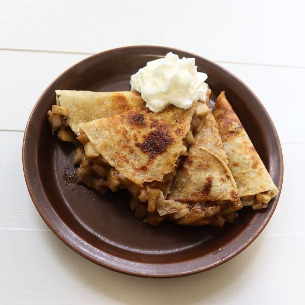 Apple pie quesadilla wedges with whipped cream on a brown plate.