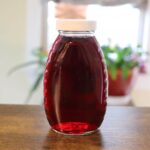 Homemade cranberry simple syrup in a clear glass bottle with a white lid on a wooden table.