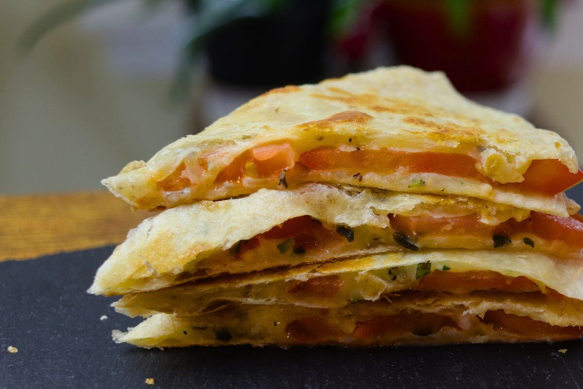 Stacked wedges of grilled cheese and tomato quesadilla with melted cheese, tomato slices, and herbs on a black board.