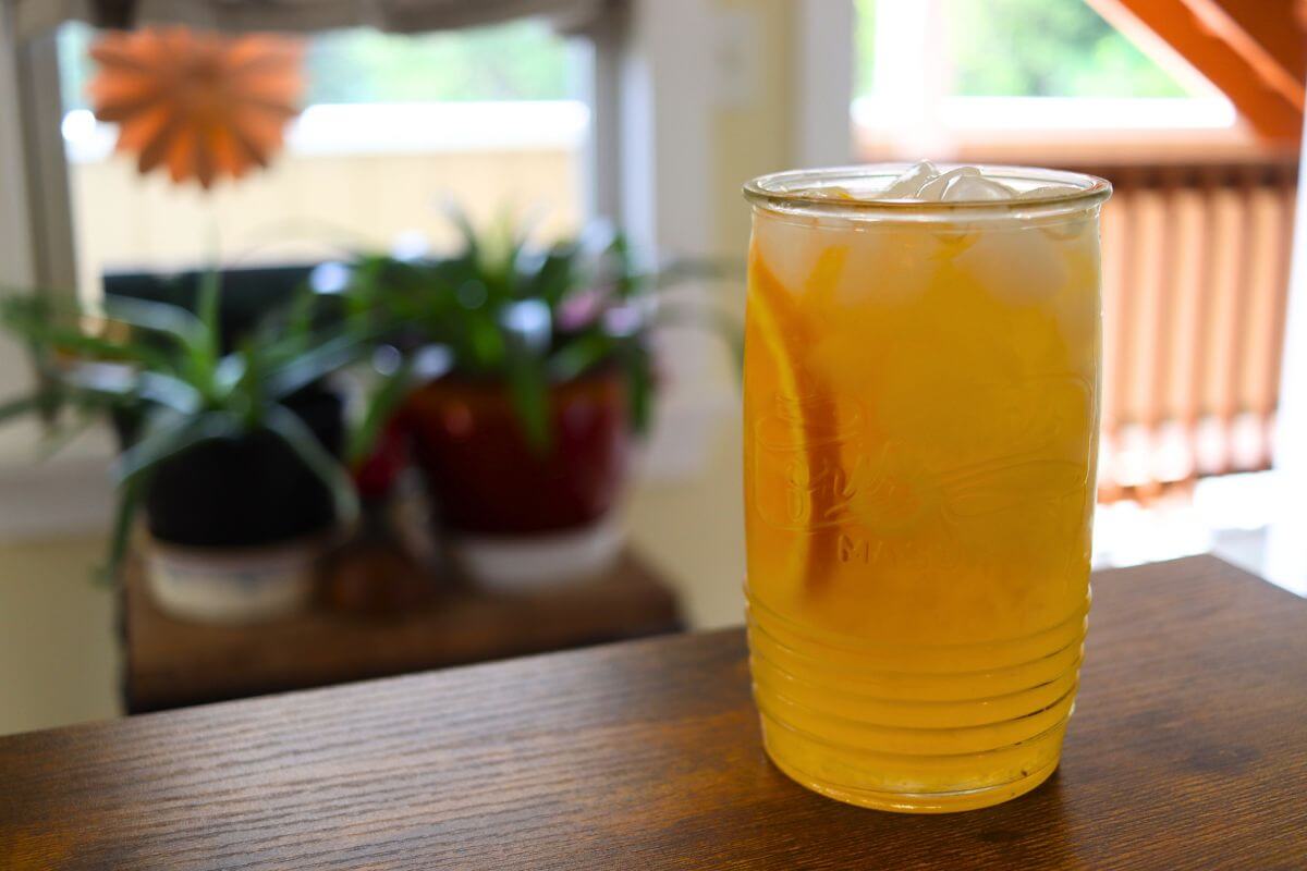 Glass of orangeade with ice on a wooden table in front of a blurred window and houseplants.