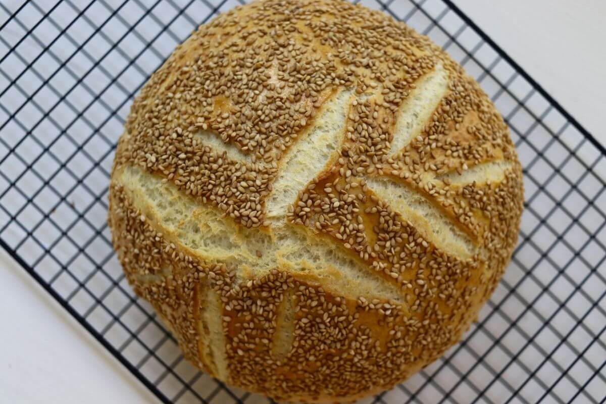 Round loaf of sesame semolina bread with decorative scoring resting on a wire cooling rack.