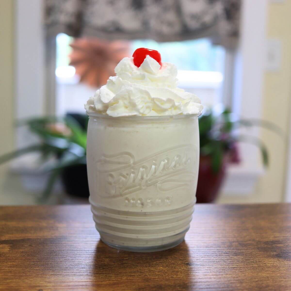 Malted milkshake in a glass with whipped cream and a maraschino cherry.