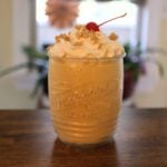 Pumpkin milkshake in a glass topped with whipped cream, graham cracker crumbs, and a maraschino cherry.