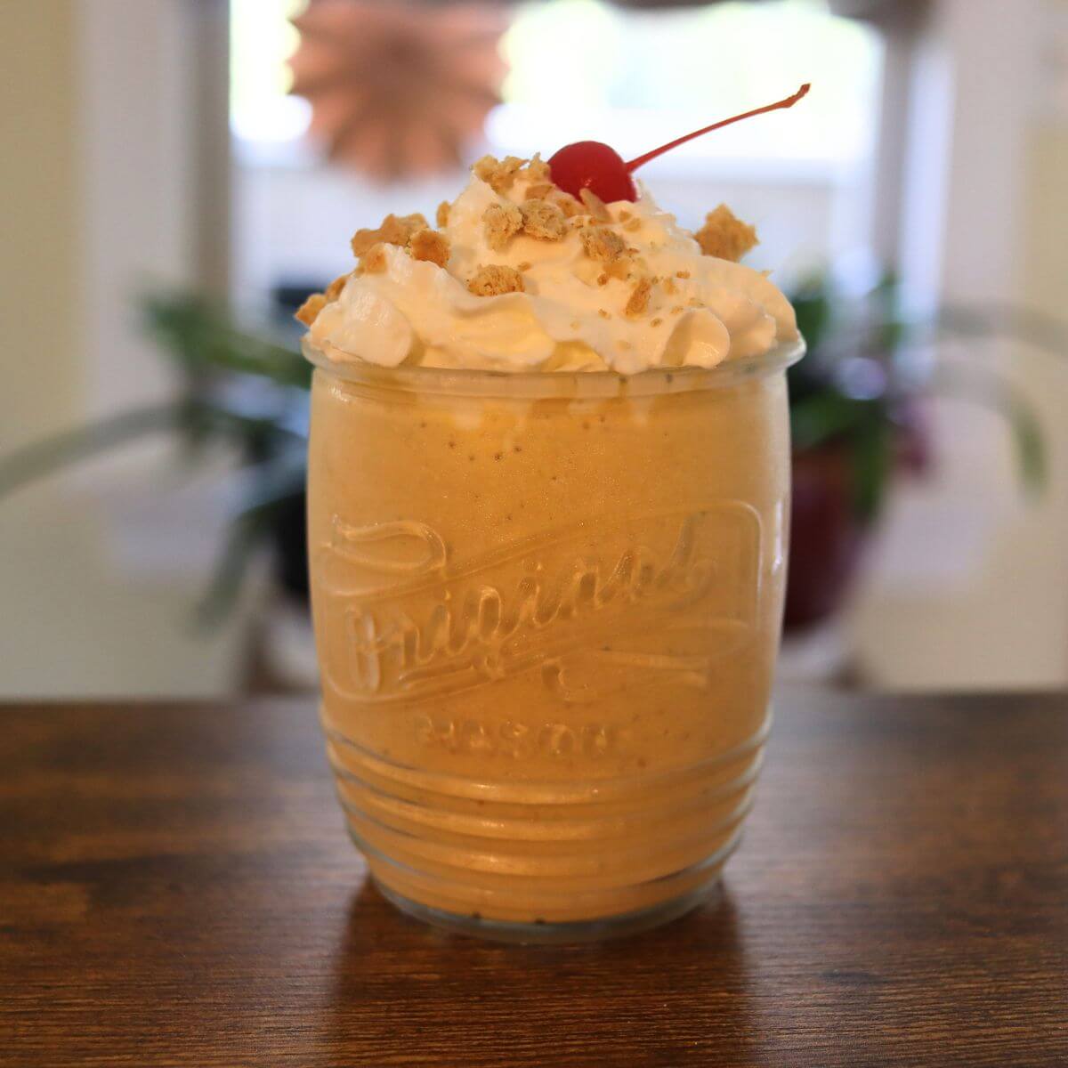 Pumpkin milkshake in a glass topped with whipped cream, graham cracker crumbs, and a maraschino cherry.