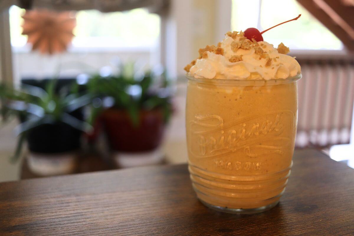 Glass filled with a pumpkin milkshake topped with whipped cream, graham cracker crumbs, and a maraschino cherry on a wooden table.