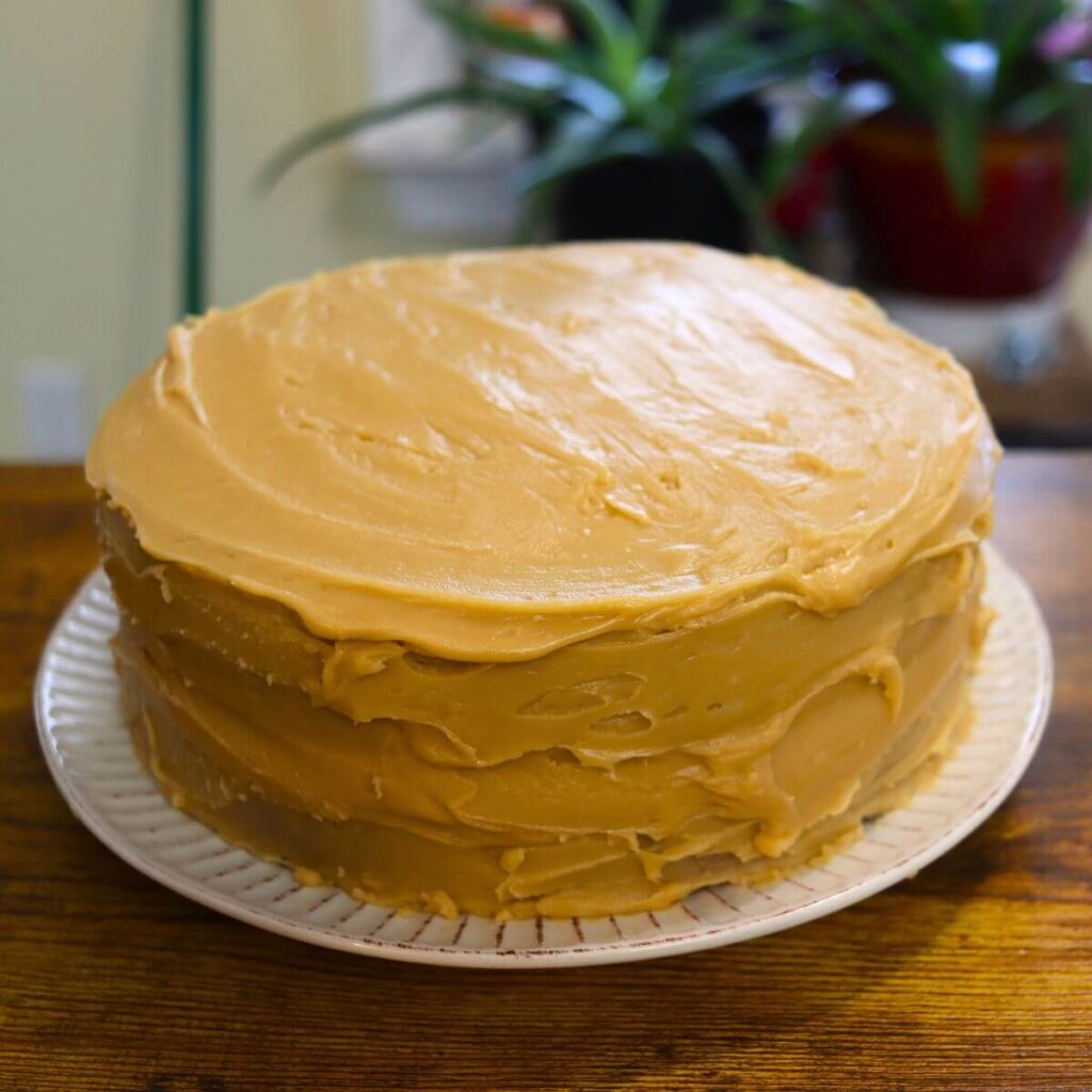 Vanilla caramel cake frosted with homemade caramel frosting on a white cake plate.