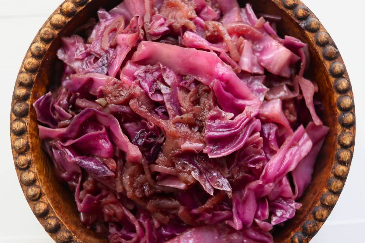 Cooked red cabbage, apples, and sliced onions piled in a round carved wooden serving bowl.