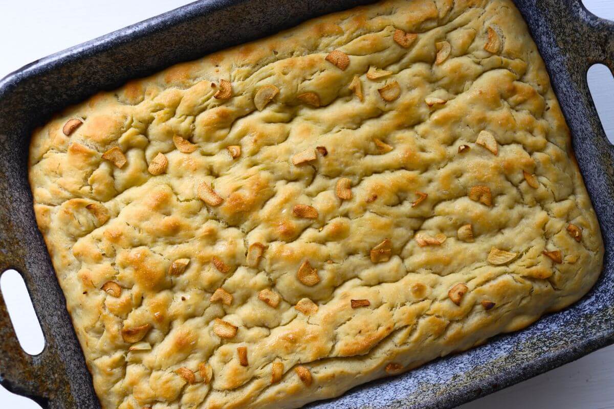 Thick garlic focaccia with golden browned areas and sliced garlic scattered across the dimpled top.