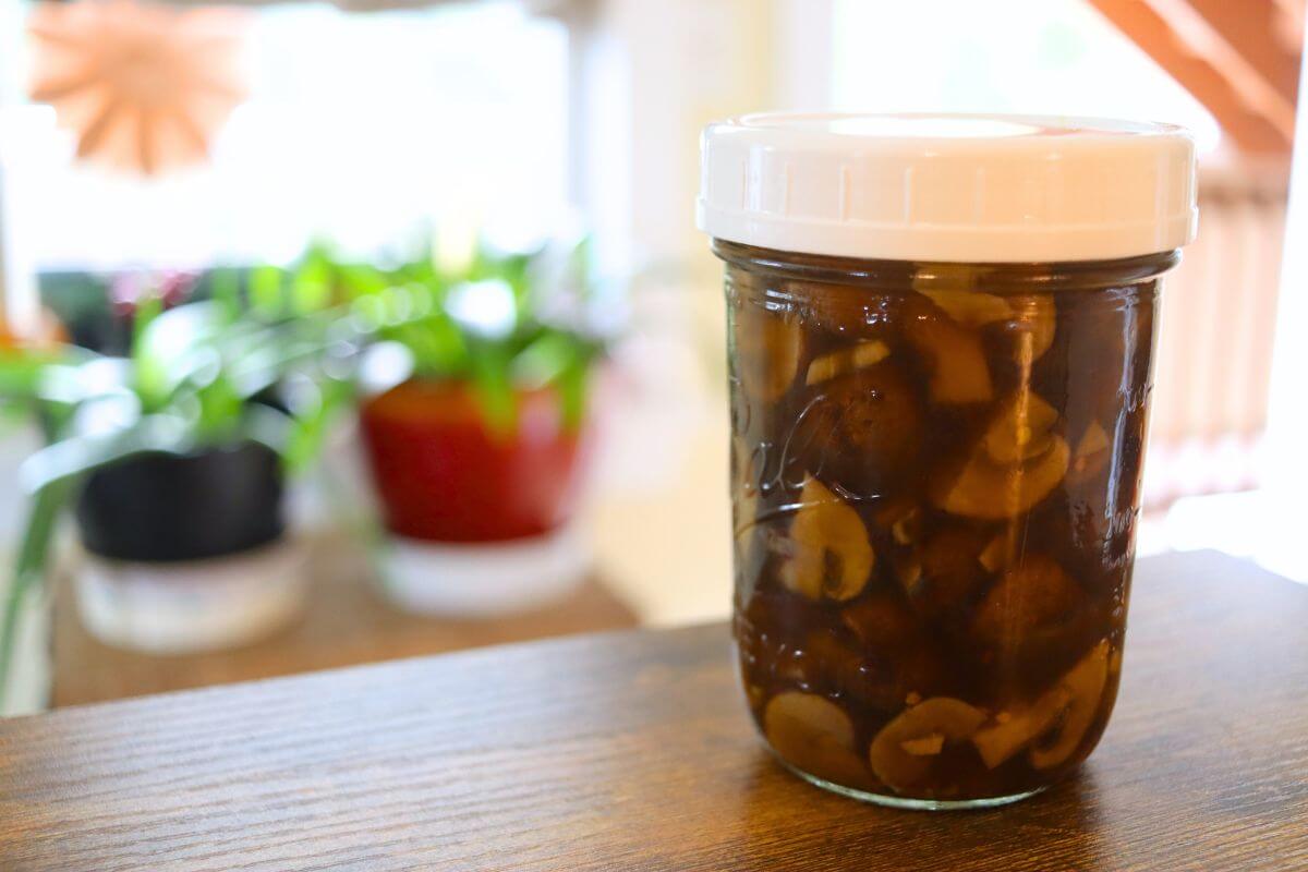 Jar of sliced pickled mushrooms in dark brine on a wooden table near a window.