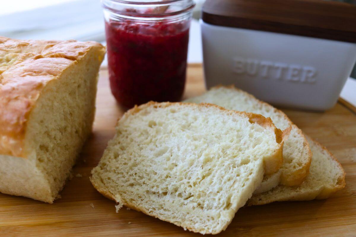 Sliced soft white sandwich bread on a wooden board with raspberry jam and a butter crock.