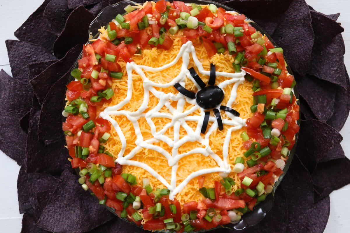 Halloween spider web dip surrounded by blue corn tortilla chips with a sour cream web and black olive spider.