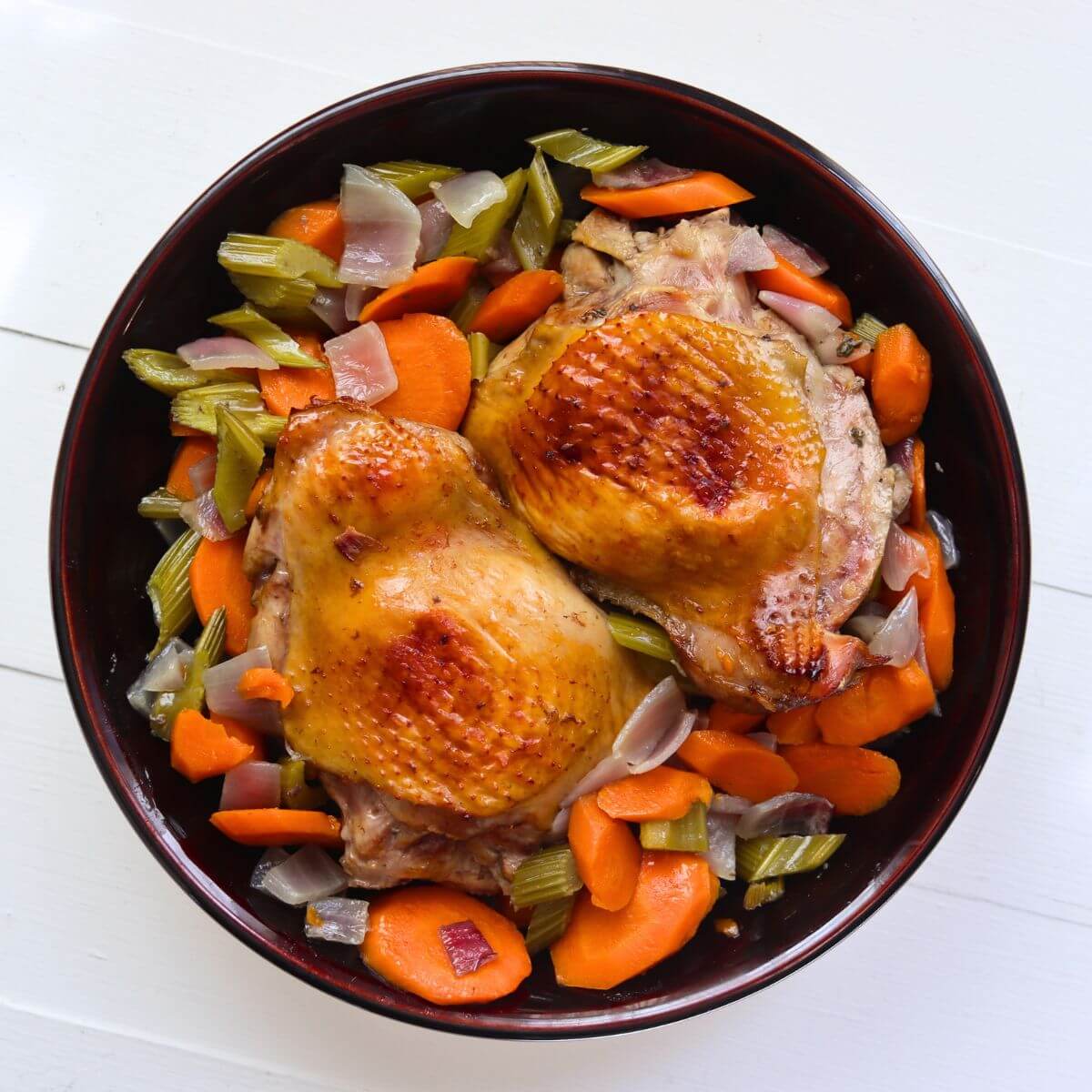 Roasted turkey thighs with golden skin nestled among cider-roasted carrots, celery, and onions in a serving bowl.